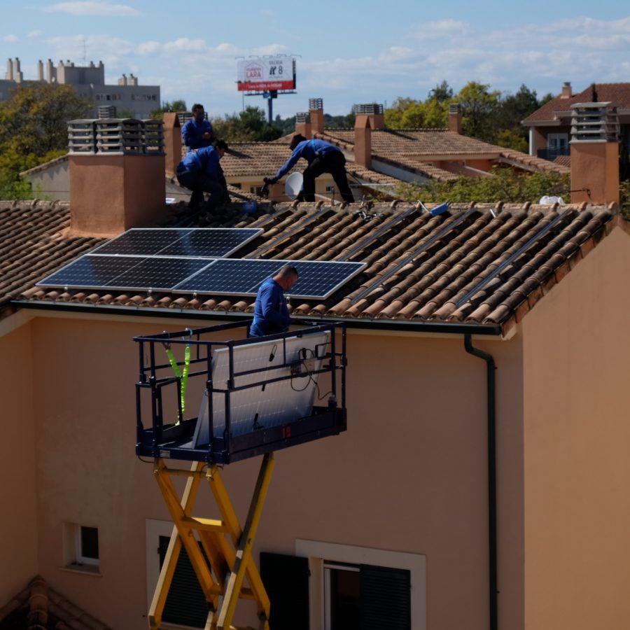 Fotografía de urbanización de casa adosadas en vista aérea donde se observan placas solares en los tejados de las casas junto a trabajadores en su instalación.